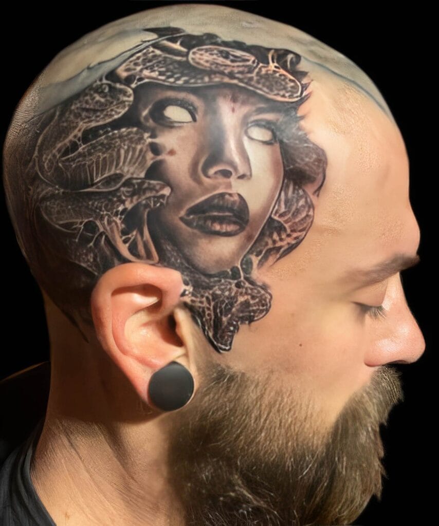 Close-up of a person’s head tattoo depicting a detailed, surreal face with snakes entwined around it. The person also has a beard and is wearing a black plug earring.