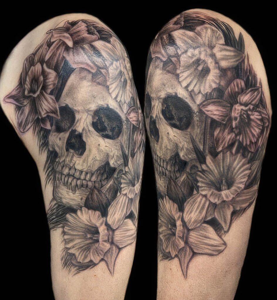 A tattoo of a skull surrounded by blooming flowers, predominantly daffodils, on an upper arm. The design is monochromatic with intricate shading.