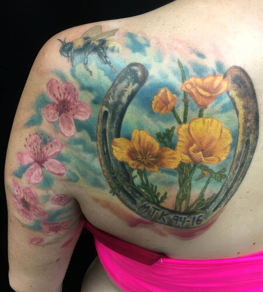 A colorful tattoo on a person's upper back featuring pink flowers, orange poppies, a horseshoe, and a bee, with the text "MTK 94-16" inscribed on the horseshoe.