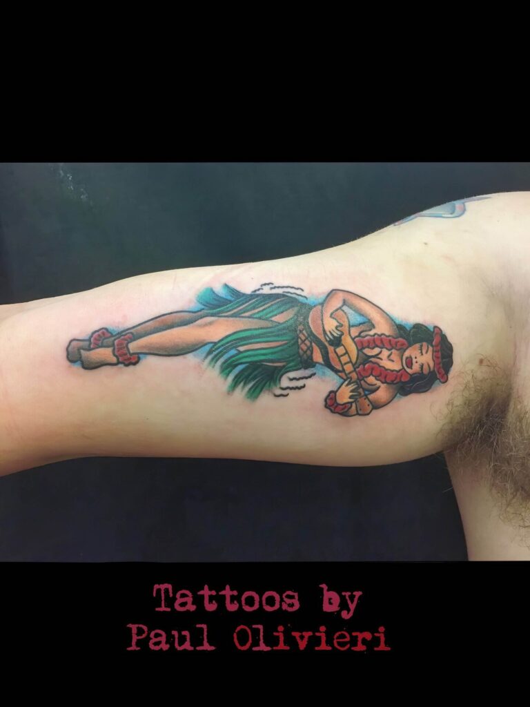 A colorful tattoo of a woman in a grass skirt playing a ukulele is inked on someone's inner arm. Below the tattoo, text reads "Tattoos by Paul Olivieri.