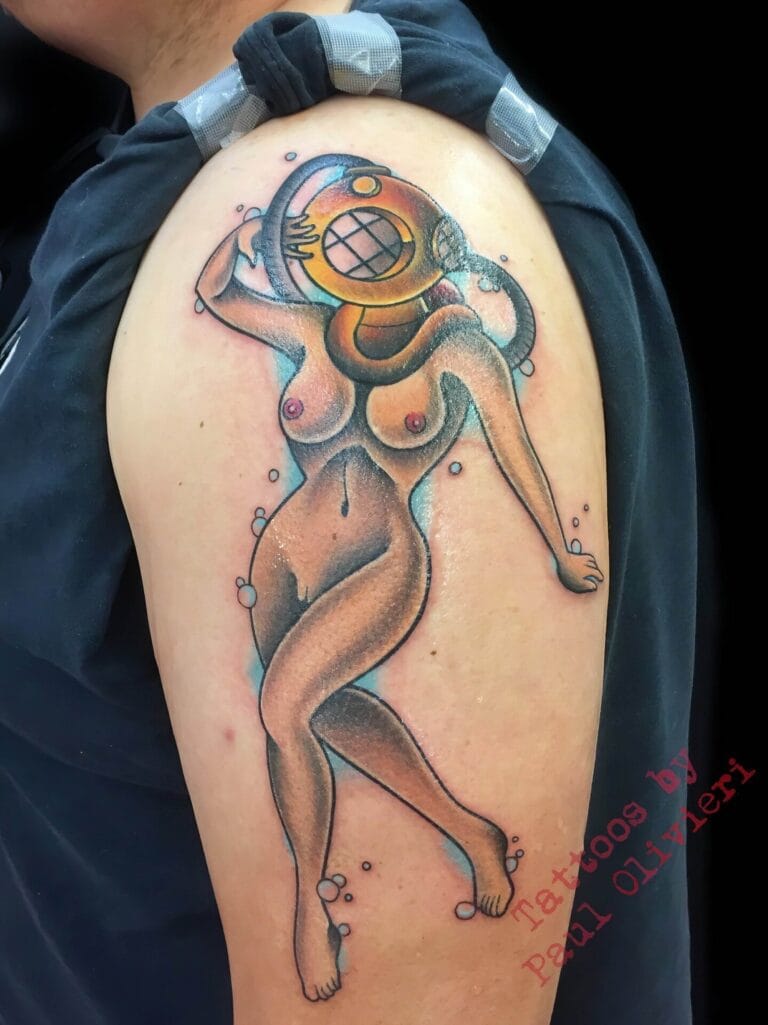 A tattoo of a stylized nude woman with a vintage diving helmet on her head, located on a person's upper arm. The image features the text "Tattoos by Paul Oliveri.
