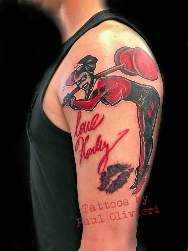 A tattoo on a person's upper arm features Harley Quinn with a hammer, "love Harley" in red text, and a black lipstick mark. The text "Tattoos by Paul Olivieri" is overlaid in red.