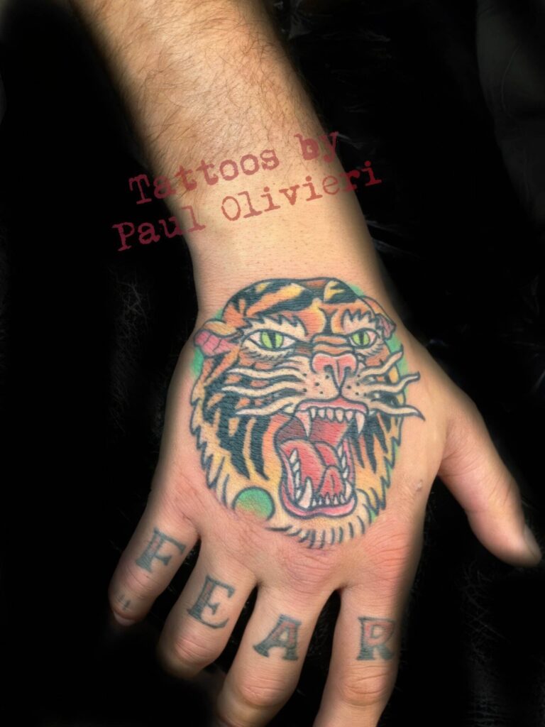 A hand tattoo features a roaring tiger head with green eyes and the word "FEAR" inked across the fingers. Text above reads "Tattoos by Paul Olivieri.