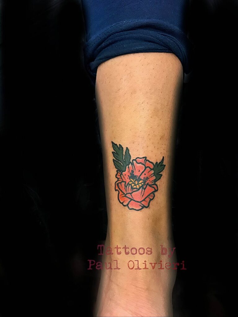 A colorful tattoo of a red flower with green leaves is displayed on a person's leg. Text in the image reads: "Tattoos by Paul Olivieri.