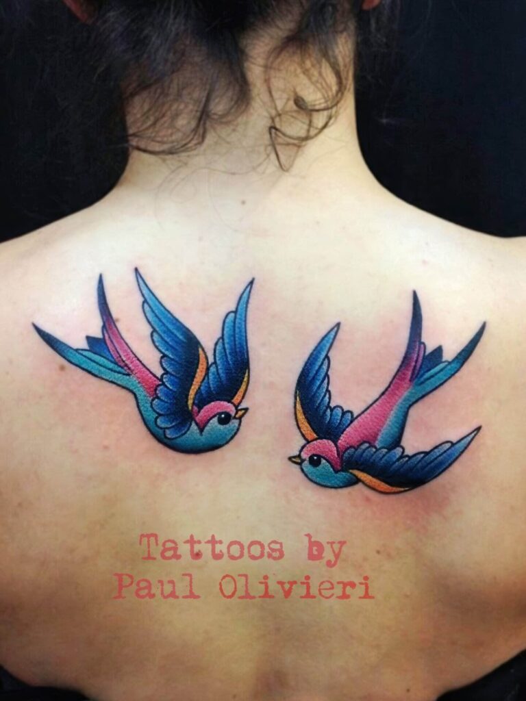 A person's upper back displays a tattoo of two colorful flying birds with blue, pink, and purple hues. Text at the bottom reads "Tattoos by Paul Olivieri.