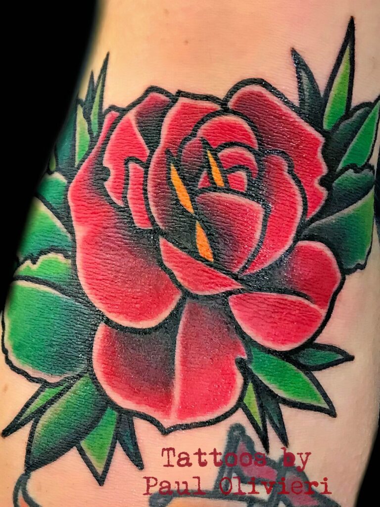 Close-up of a vibrant red rose tattoo with green leaves, prominently featuring the text "Tattoos by Paul Olivieri" at the bottom.