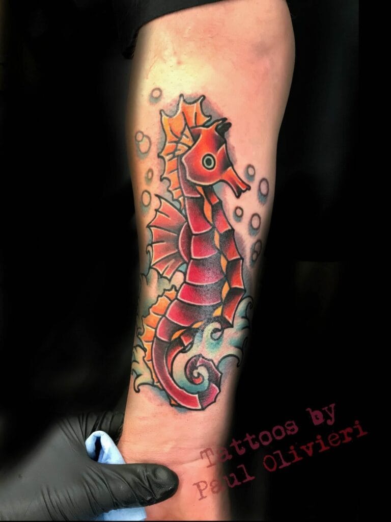 A colorful seahorse tattoo on a person's forearm with bubbles surrounding it. A gloved hand is seen holding a cloth at the bottom of the arm. The text "Tattoos by Paul Olivieri" is in the corner.