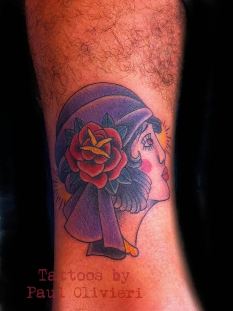 A tattoo of a woman's profile with a large red rose on her head, featuring a purple headscarf, detailed shading, and vibrant colors. Text below reads, "Tattoos by Paul Olivieri.