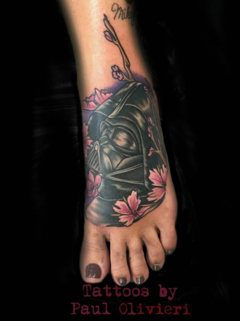 A foot tattoo depicting a helmeted figure surrounded by pink flowers, with text at the bottom reading "Tattoos by Paul Olivieri.