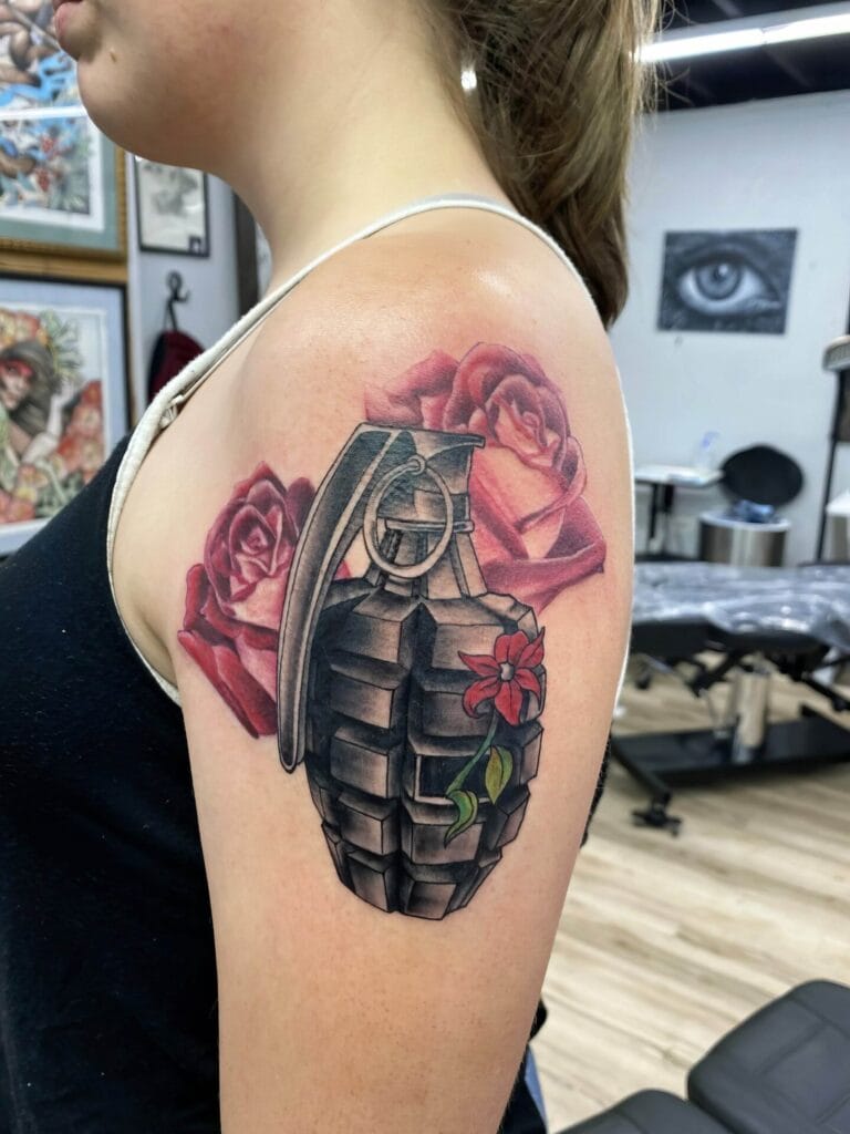 Upper arm tattoo featuring a black grenade with a red flower and green leaves in front of red roses on a person's shoulder.