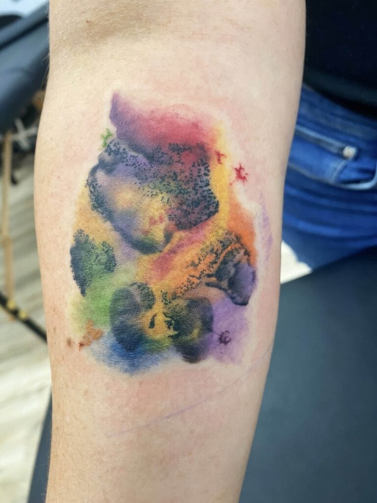 A forearm with a colorful abstract tattoo featuring splashes of purple, yellow, orange, and green ink.