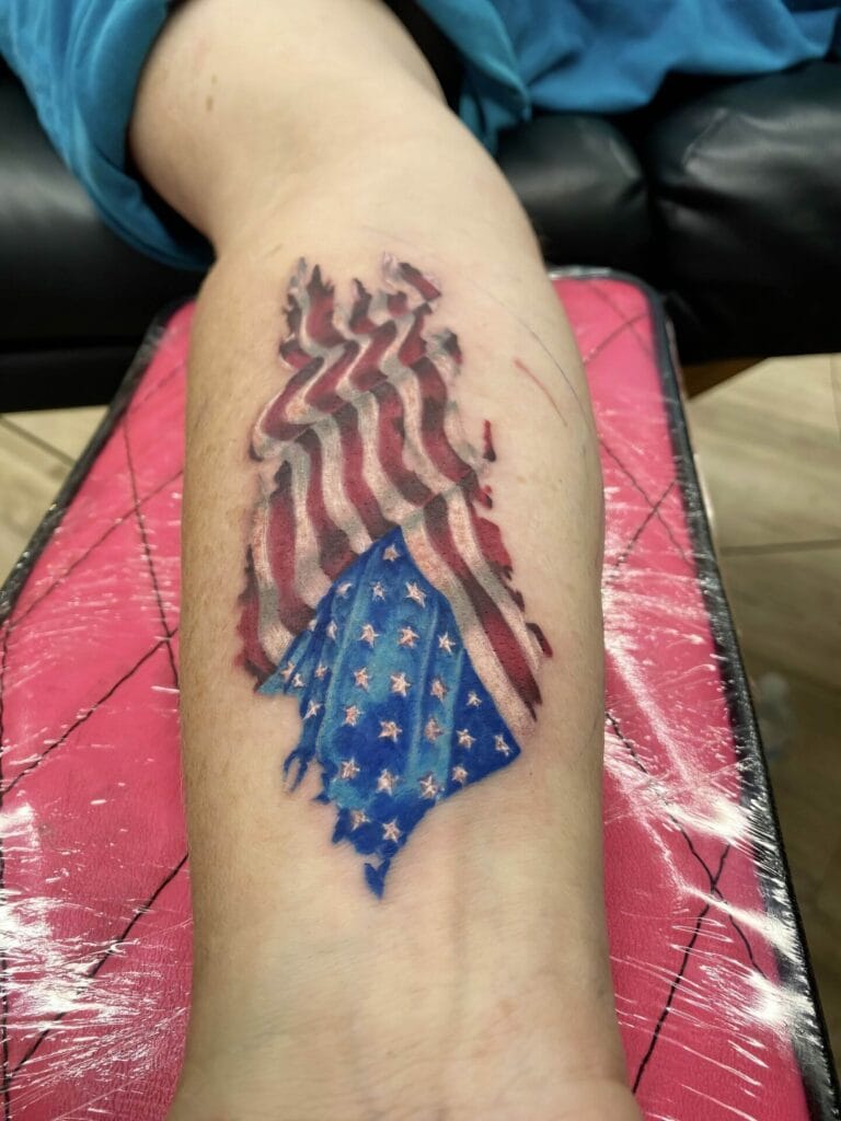 A person's forearm with a tattoo of an American flag, where the top part of the flag appears to be burning away. The tattoo shows vivid red, white, and blue colors.