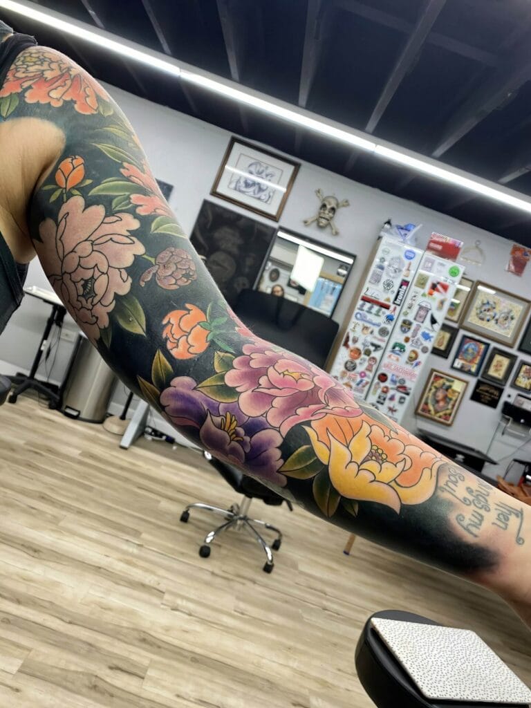 A tattooed arm with colorful floral designs including peonies and other flowers, showcased in a tattoo studio.