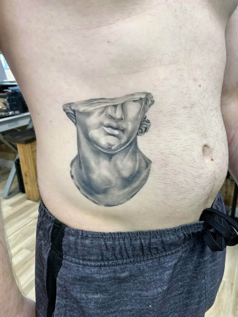 A person with a tattoo on their side featuring a bust of a classical statue with the eyes obscured, wearing gray sweatpants.