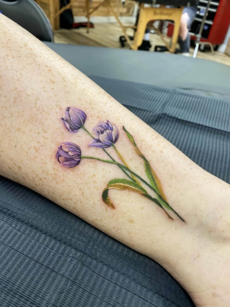 A person has a tattoo of three purple tulips with green leaves on their forearm against a neutral background.