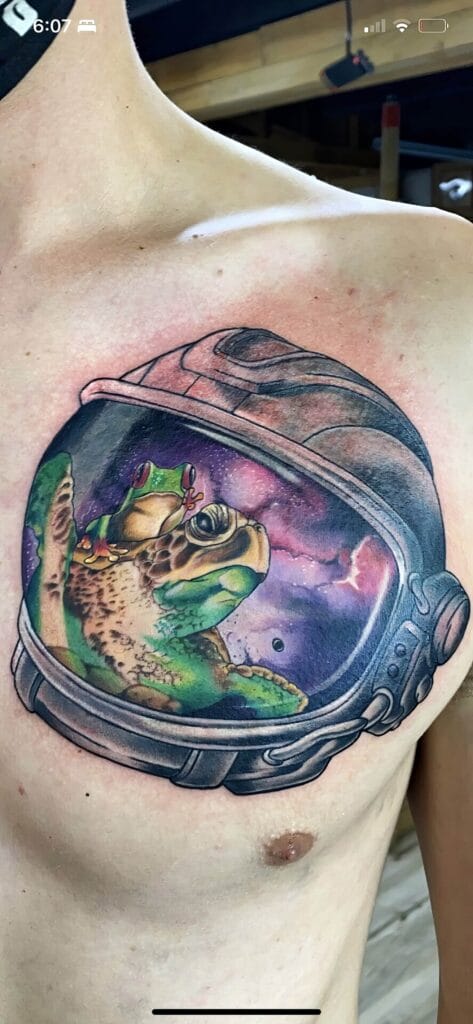 A tattoo on a person's chest depicting a frog in an astronaut helmet with a colorful galaxy background.