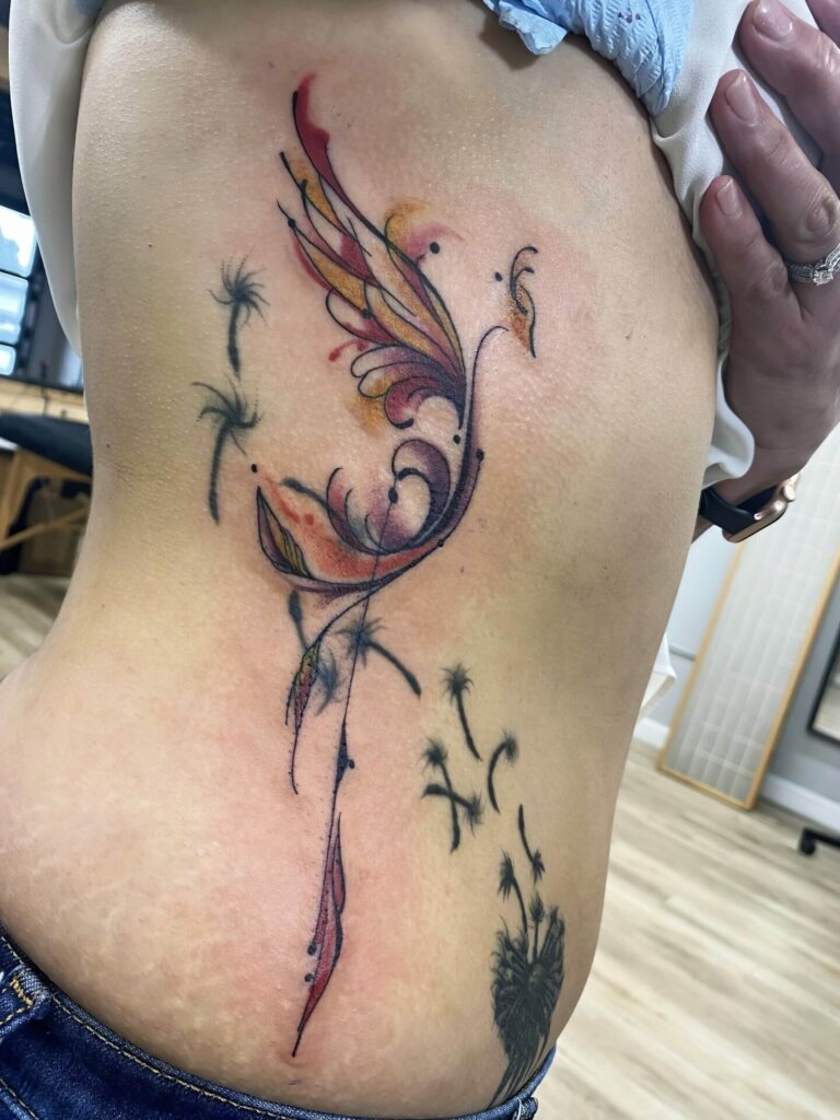 A colorful tattoo of a stylized phoenix with swirling lines and feather details on a person's side torso, surrounded by scattered dandelion seeds.