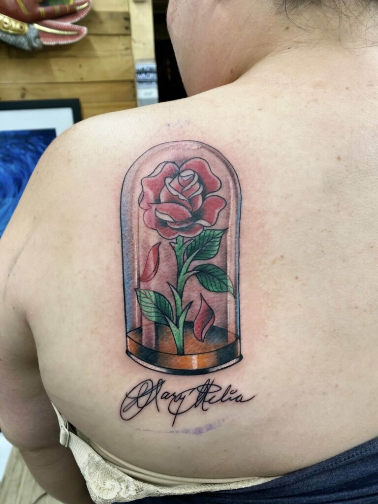 Tattoo of a rose enclosed in a glass dome with the name "Mary Alia" written underneath, located on a person's upper back.