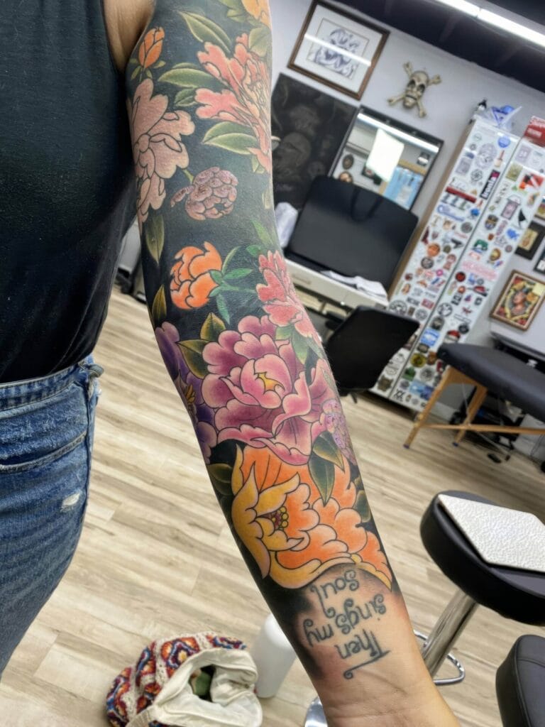 A person with a colorful floral tattoo sleeve on their arm, featuring various flowers and leaves, stands in a tattoo studio. The text "then sings my soul" is visible among the flowers.