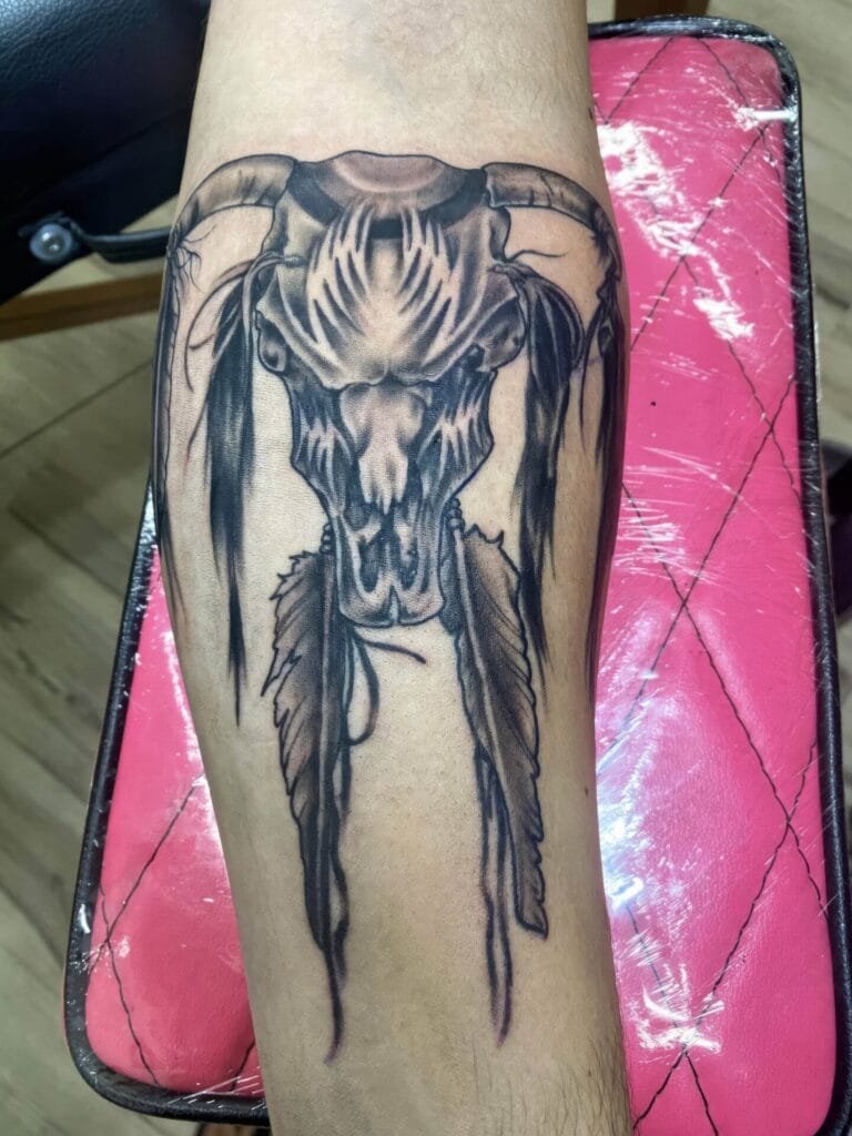 A black and grey tattoo on an arm features a skull with horns, adorned with hanging feathers. The tattoo is shown on a red cushioned surface.