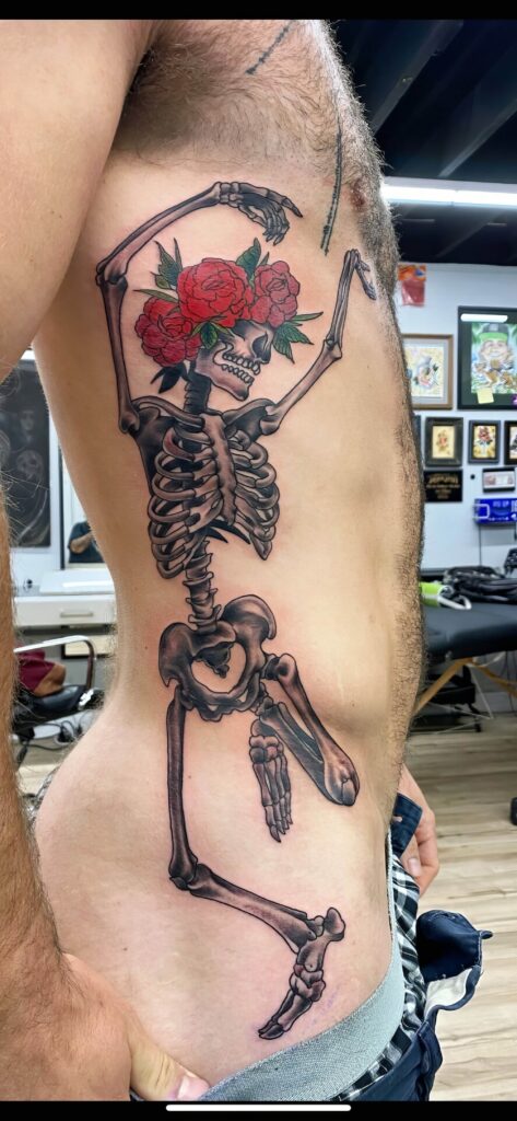 A tattoo of a dancing skeleton with red roses for a face is inked on a person’s side torso. The detailed art extends from the ribcage to the hip.