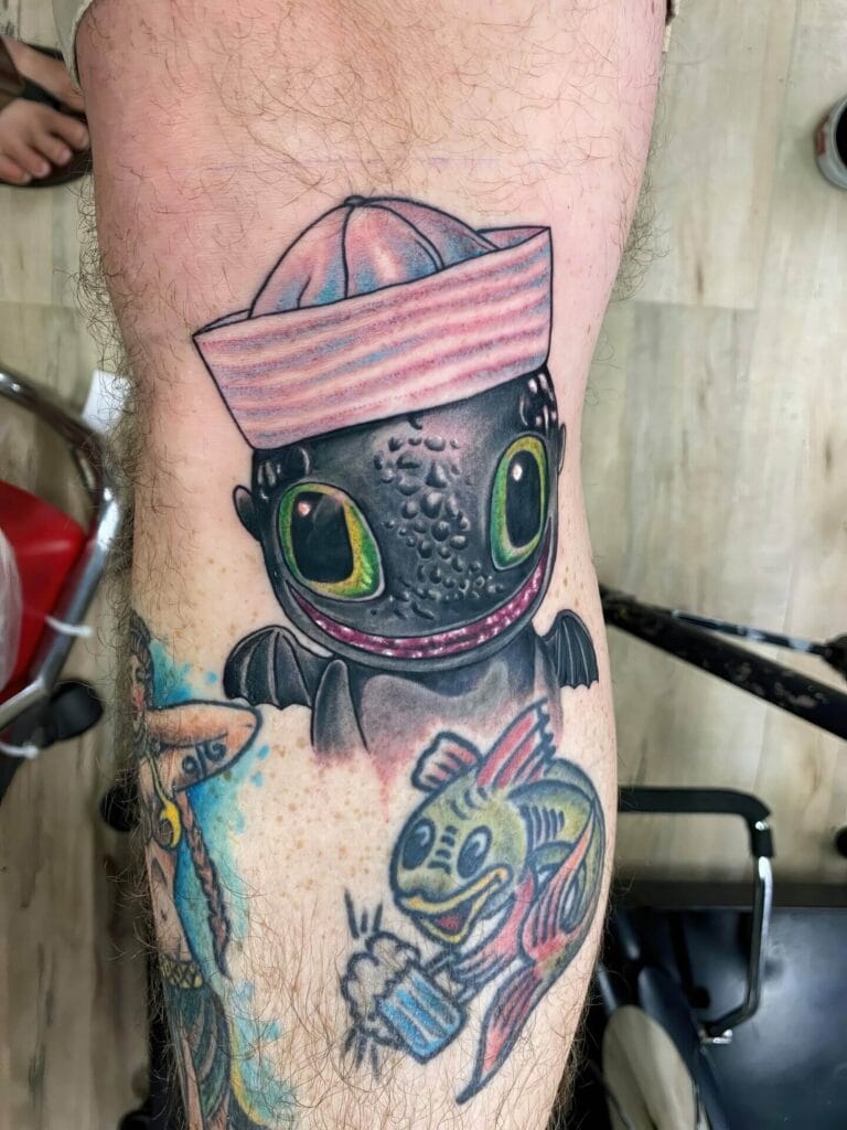 A leg featuring a tattoo of a cartoonish black dragon wearing a pink sailor hat, above a tattoo of a colorful fish holding a beer in a frosty mug.