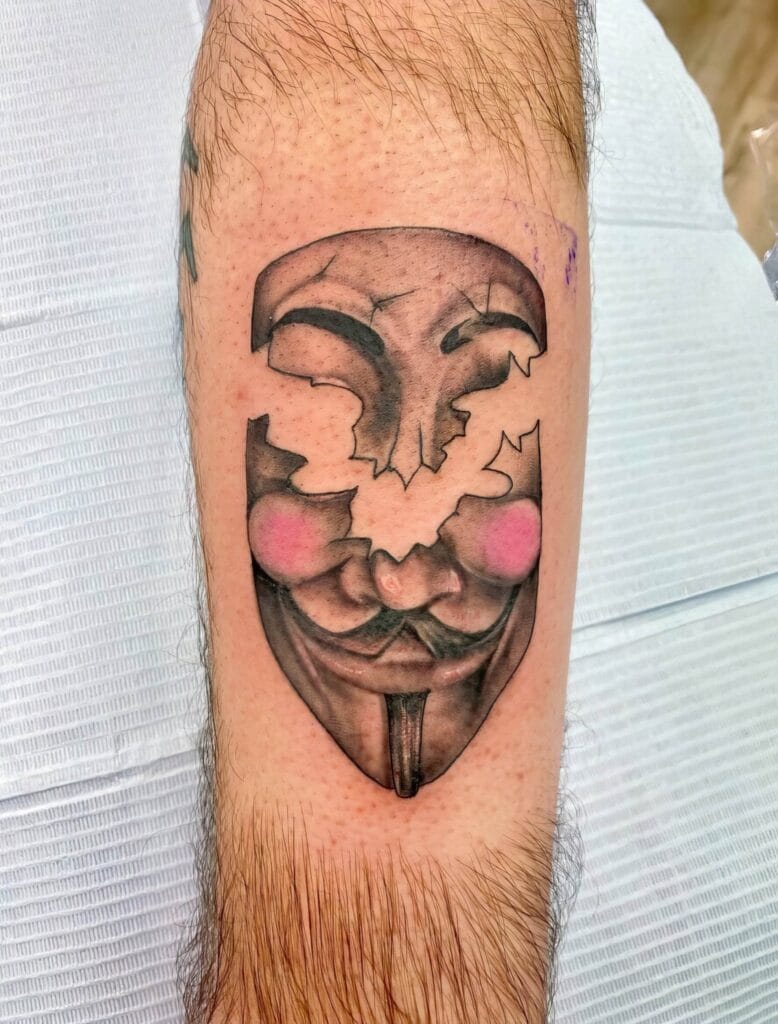 A forearm tattoo featuring a stylized mask with red cheeks, reminiscent of the Guy Fawkes mask from "V for Vendetta," against a white background.