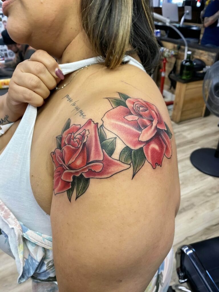 A person displays a detailed tattoo of two large red roses with green leaves on their upper arm and shoulder in a tattoo parlor.