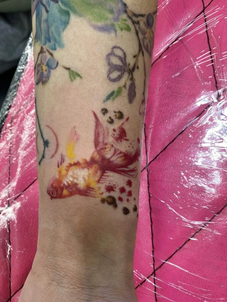 A forearm with a colorful tattoo of a fish and flowers displayed on a pink background.