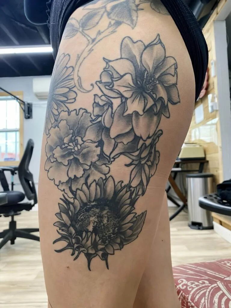 Black and gray floral tattoo featuring a sunflower, roses, and other flowers on thigh.