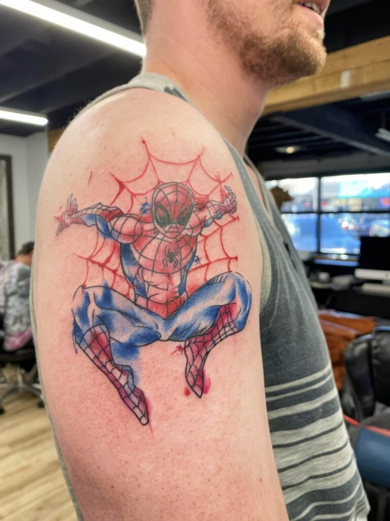 A person with a Spider-Man tattoo on their upper arm, detailed with a web background, standing in an indoor setting.