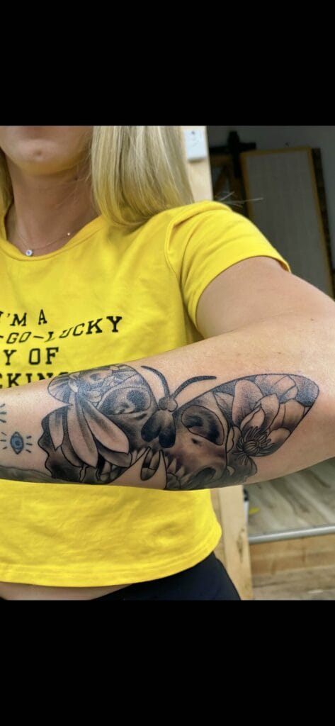 A person with blonde hair is wearing a yellow T-shirt and showcasing a detailed black and grey tattoo of a skull, flowers, and a bee on their forearm.