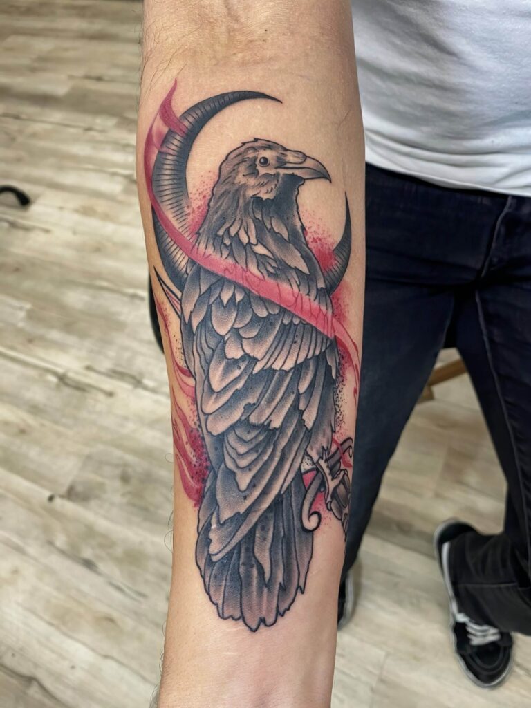 A tattoo of a raven with stylized feathers and red accents on a person's forearm.