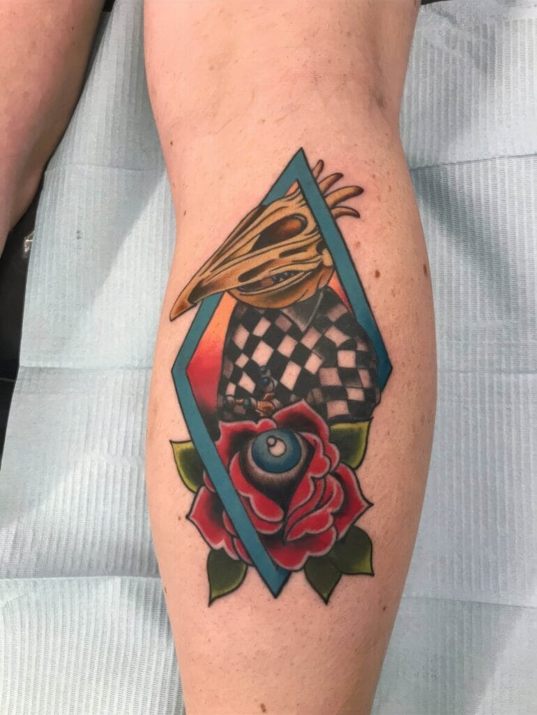 A leg tattoo featuring a bird skull, a checkered pattern, an eye, and a red rose, framed within a blue diamond shape.