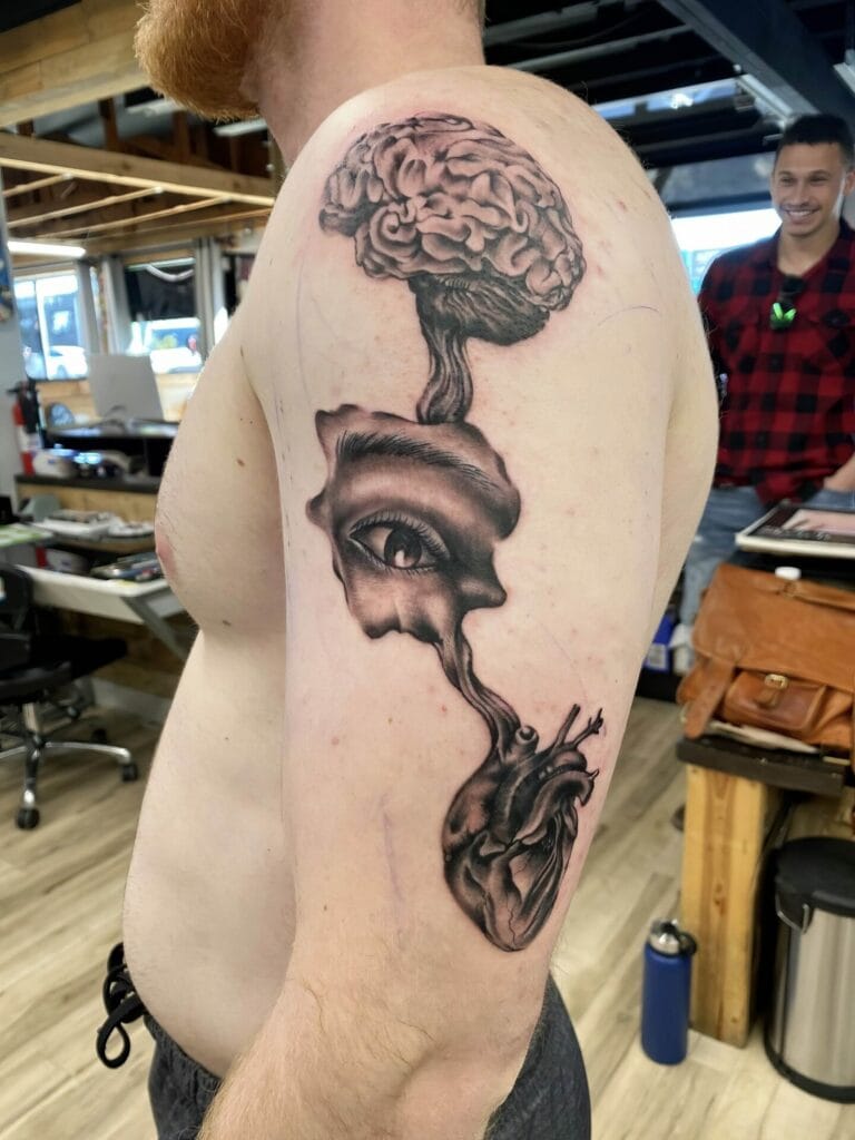 A man with a new tattoo on his upper arm featuring a brain, eye, and heart, connected by abstract designs.