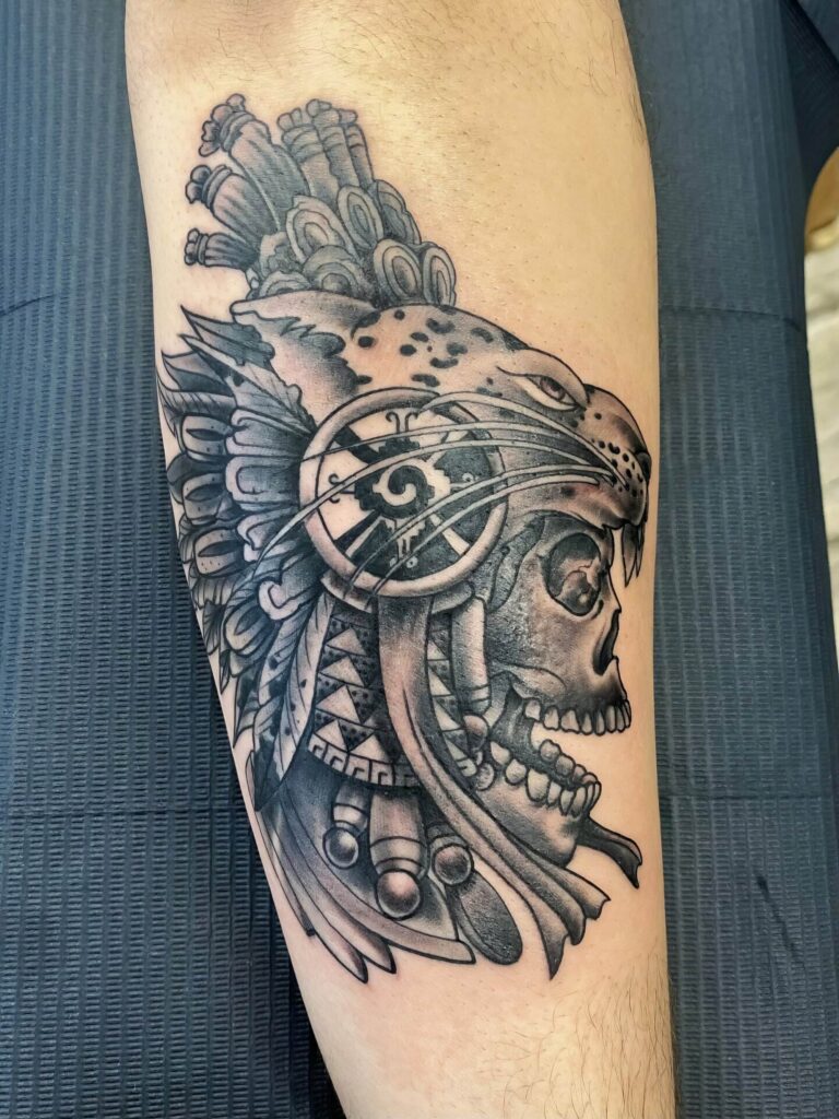 A detailed black and grey tattoo on a forearm, depicting a fierce jaguar headdress worn by a skull with elaborate tribal designs.