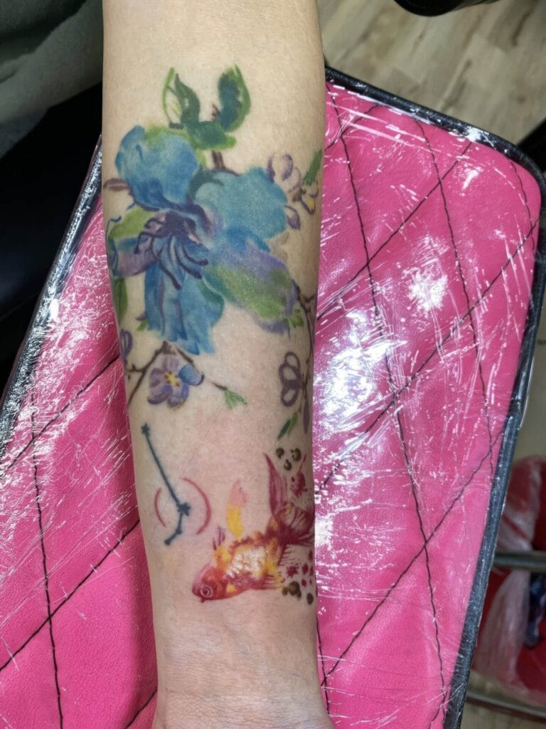 A forearm tattoo featuring a blue flower and a pink fish, with the skin underneath covered in plastic wrap, presumably to protect the new tattoo.