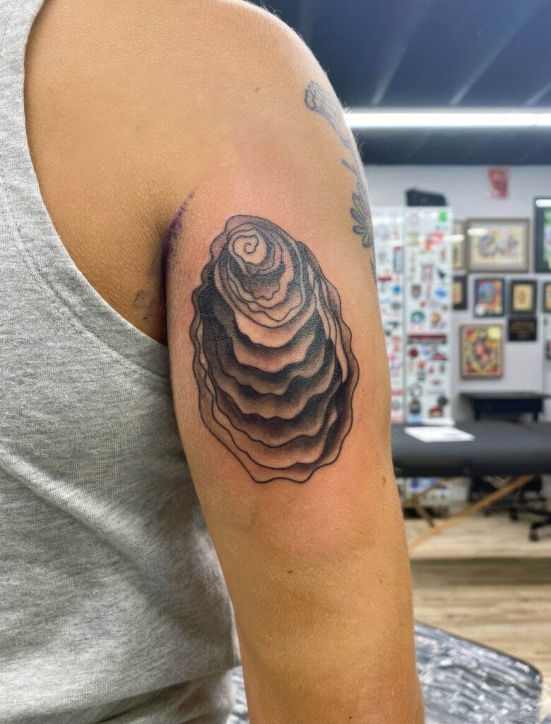 A person with a freshly inked black spiral tattoo on their upper arm, wearing a gray sleeveless shirt inside a tattoo studio.