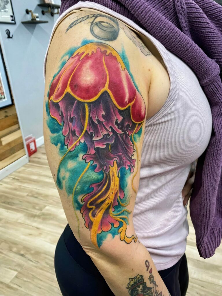 A person with a colorful jellyfish tattoo on their upper arm, wearing a sleeveless shirt and a sweater draped over their shoulder.