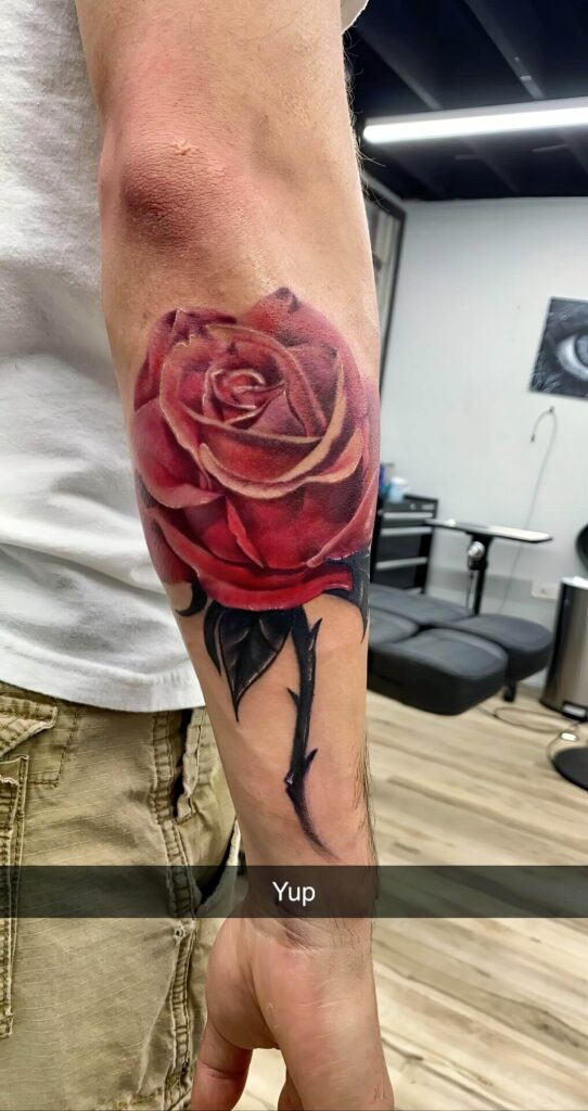 A person with a large, detailed tattoo of a red rose with a black stem on their forearm, standing in what appears to be a tattoo studio. A "Yup" caption overlays the image.