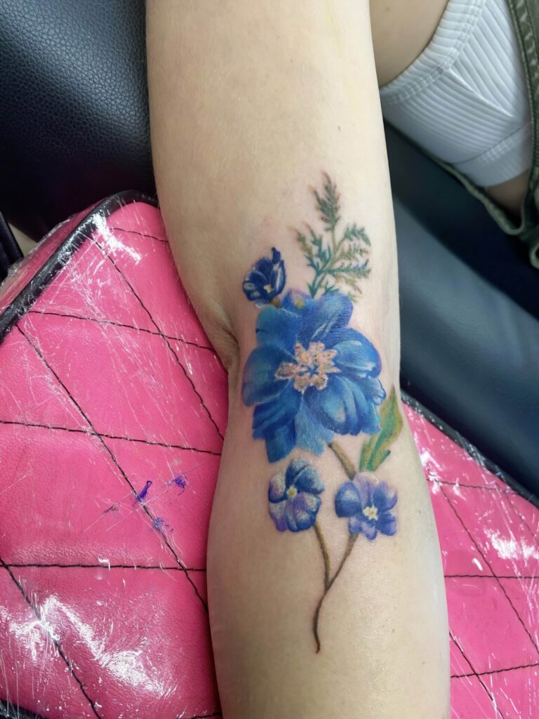 A person displays a tattoo of blue flowers with green leaves on their forearm. The tattoo is new as some redness is visible near the design.