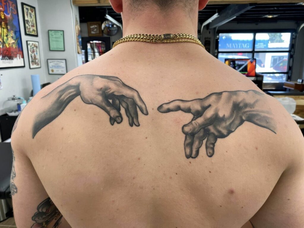 A person's back with tattoos depicting the hands from Michelangelo's "The Creation of Adam"; one on each shoulder blade.