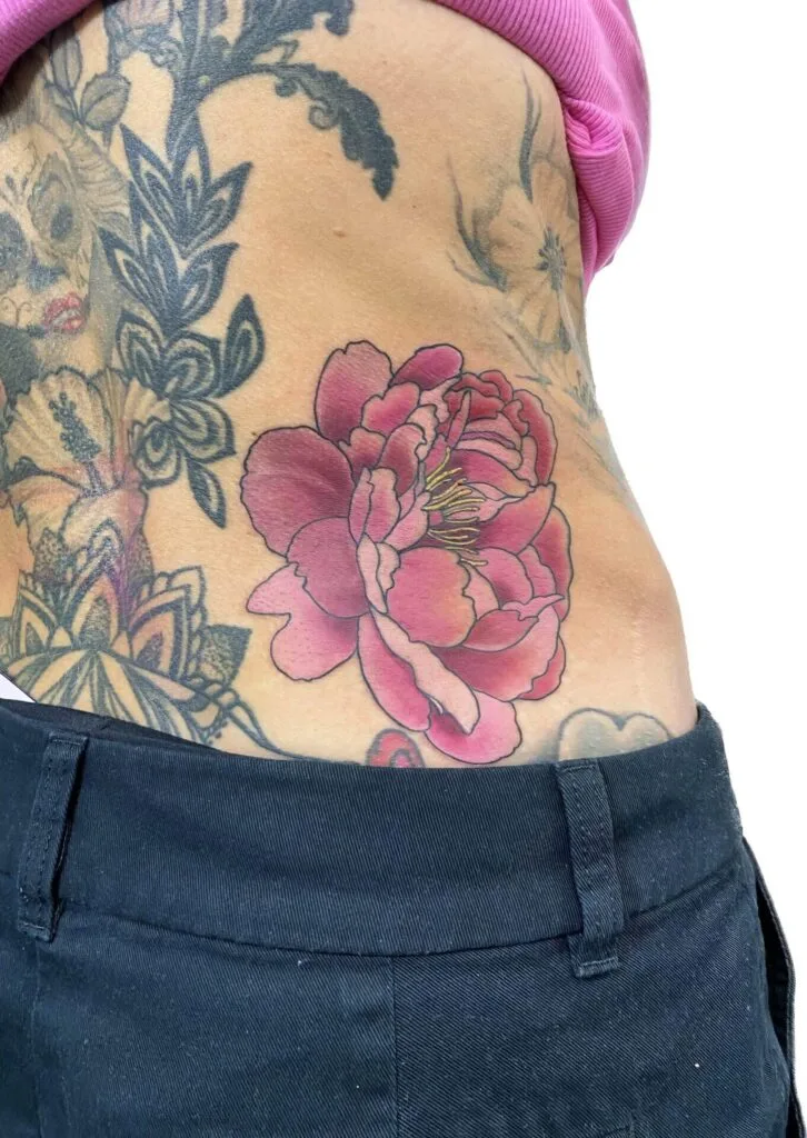 Pink and red peony tattoo on persons side waist, with other black ink tattoos nearby.