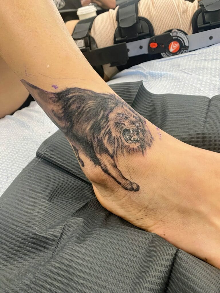 A tattoo of a roaring lion is on a person's lower leg and ankle. The person is lying on a black mat.