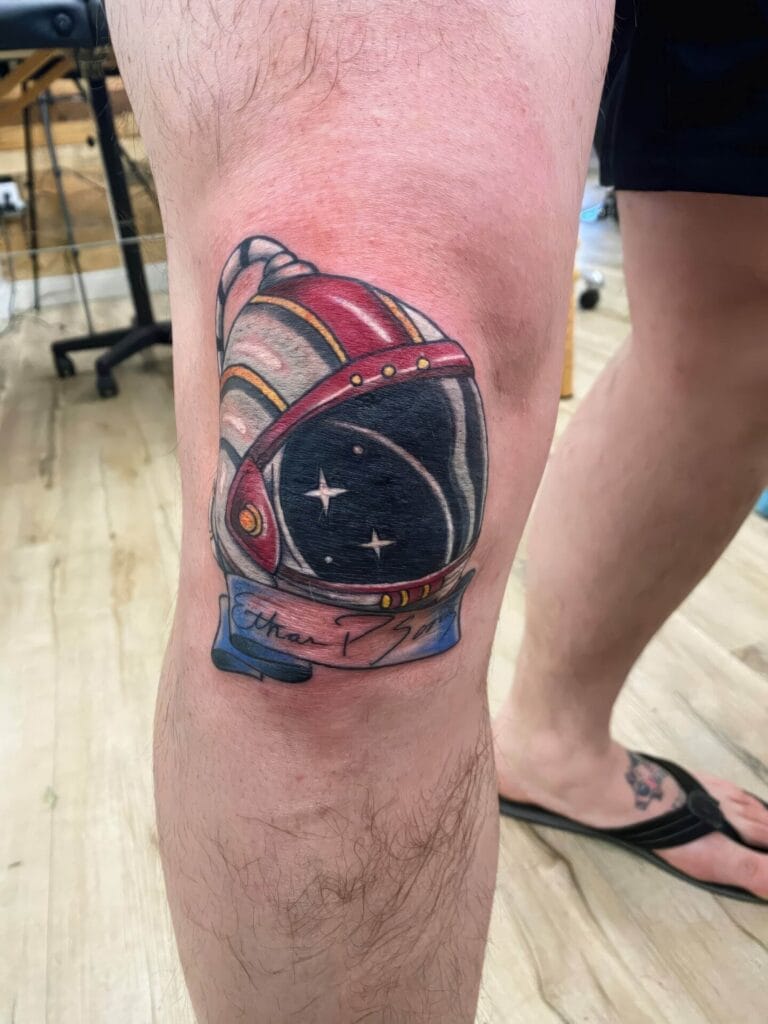 A colorful tattoo of an astronaut helmet with a banner on a person's knee, showcasing a space-themed design. The background setting appears to be indoors.