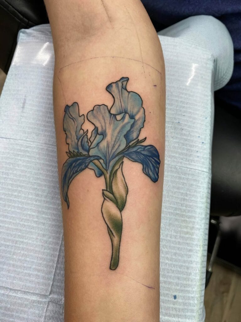 A close-up of a forearm with a blue and green flower tattoo design. The arm is resting on a white and blue textured surface.