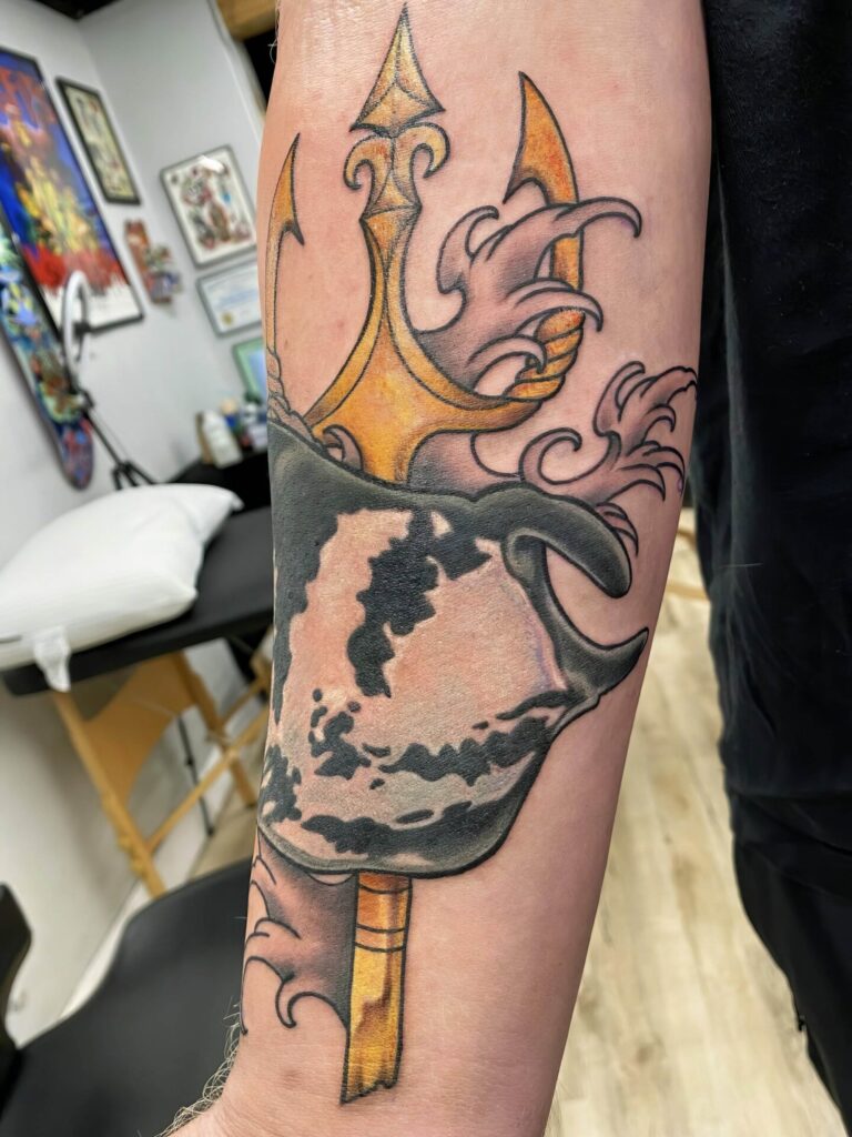 Forearm tattoo featuring a trident with decorative swirls and a helmeted face, rendered in vibrant colors. Tattoo studio background with tattoo equipment visible.