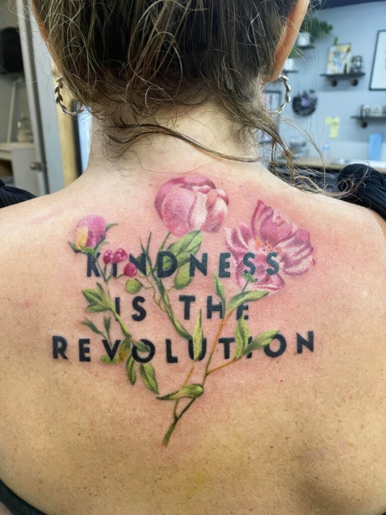 A tattoo on a person's upper back features pink flowers with green stems and leaves surrounding the text "KINDNESS IS THE REVOLUTION.