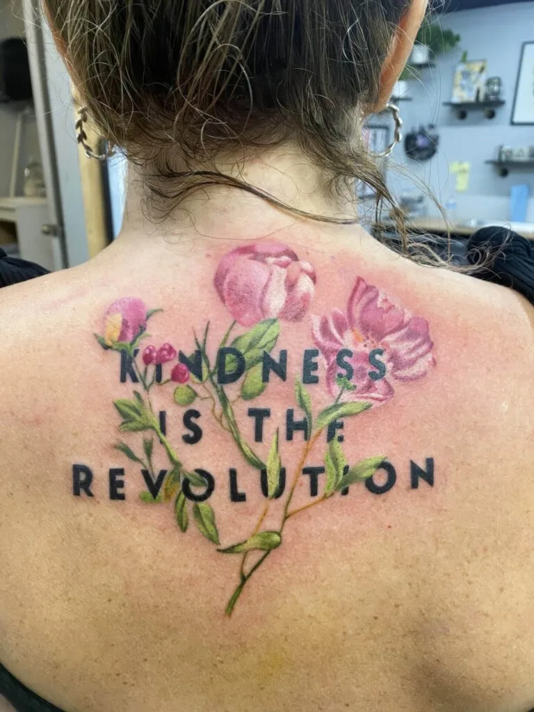 A tattoo on a person's upper back features pink flowers with green stems and leaves surrounding the text "KINDNESS IS THE REVOLUTION.