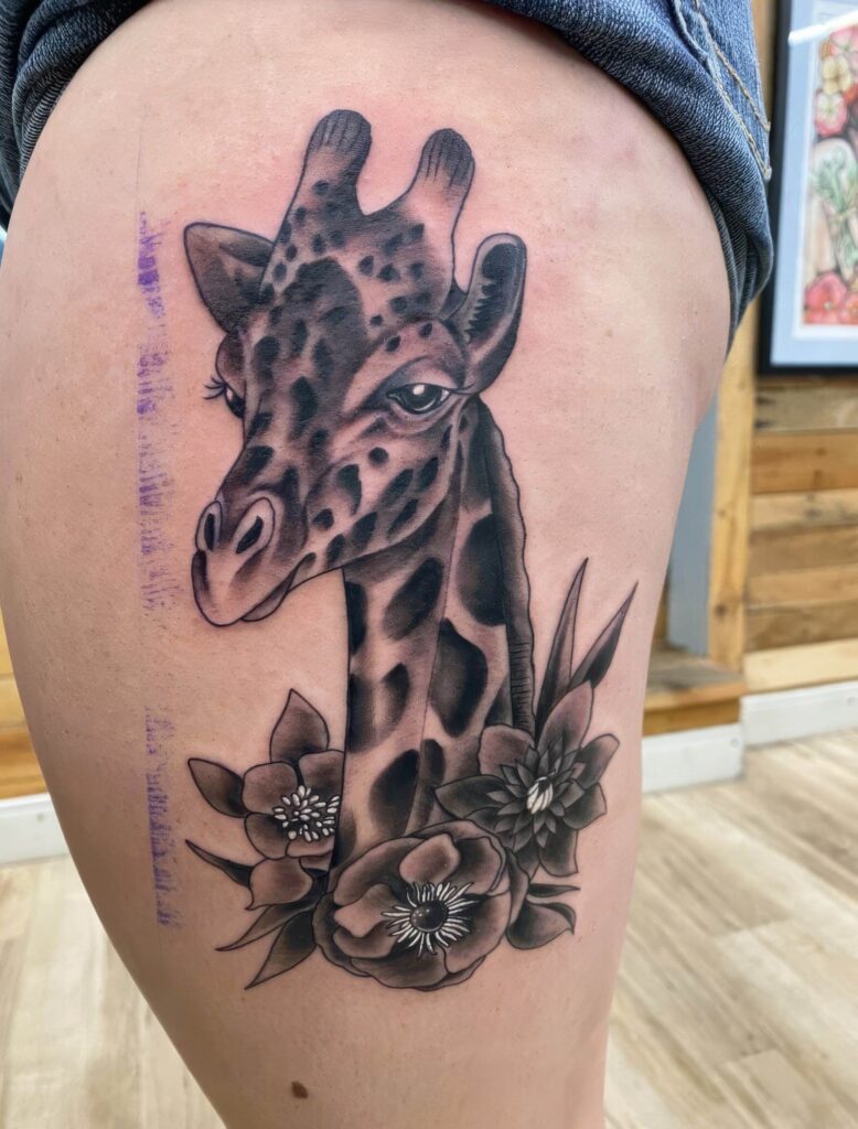 A detailed tattoo of a giraffe with flowers around its neck is seen on a person's thigh. The design features dark shading and intricate details in black and grey tones.